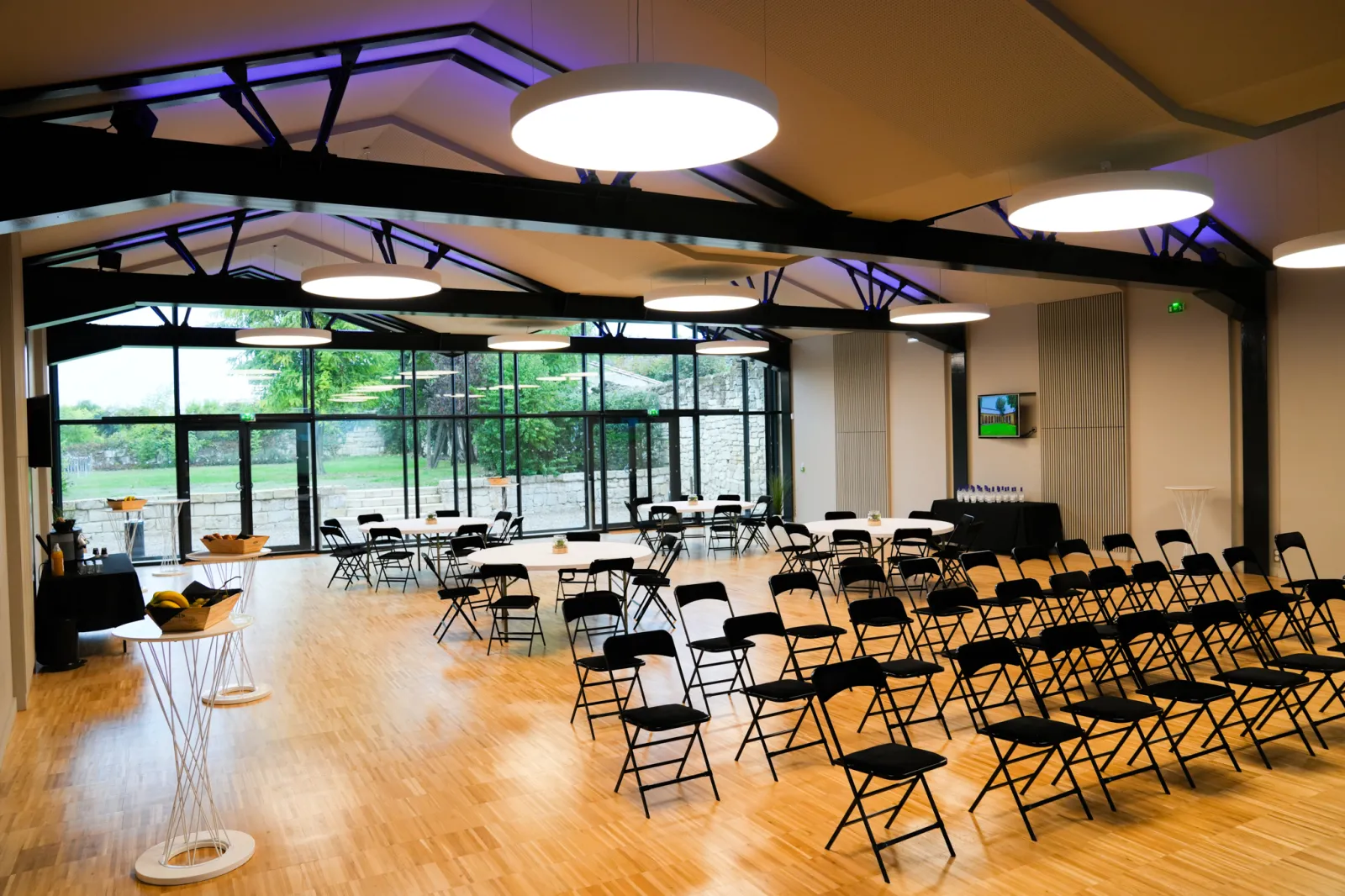 Modern event space with rows of black folding chairs on wooden flooring, large windows overlooking green landscape, round tables with gold accents, contemporary ceiling lighting, and stage area in Les Cordeliers, Bordeaux.