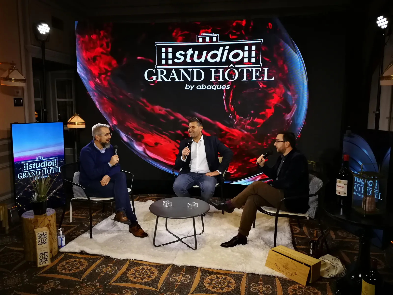 Three men in business attire having a panel discussion on stage at the Studio Grand Hotel by Abaques in Bordeaux, with a large illuminated backdrop displaying red and blue abstract visuals and the hotel's logo.