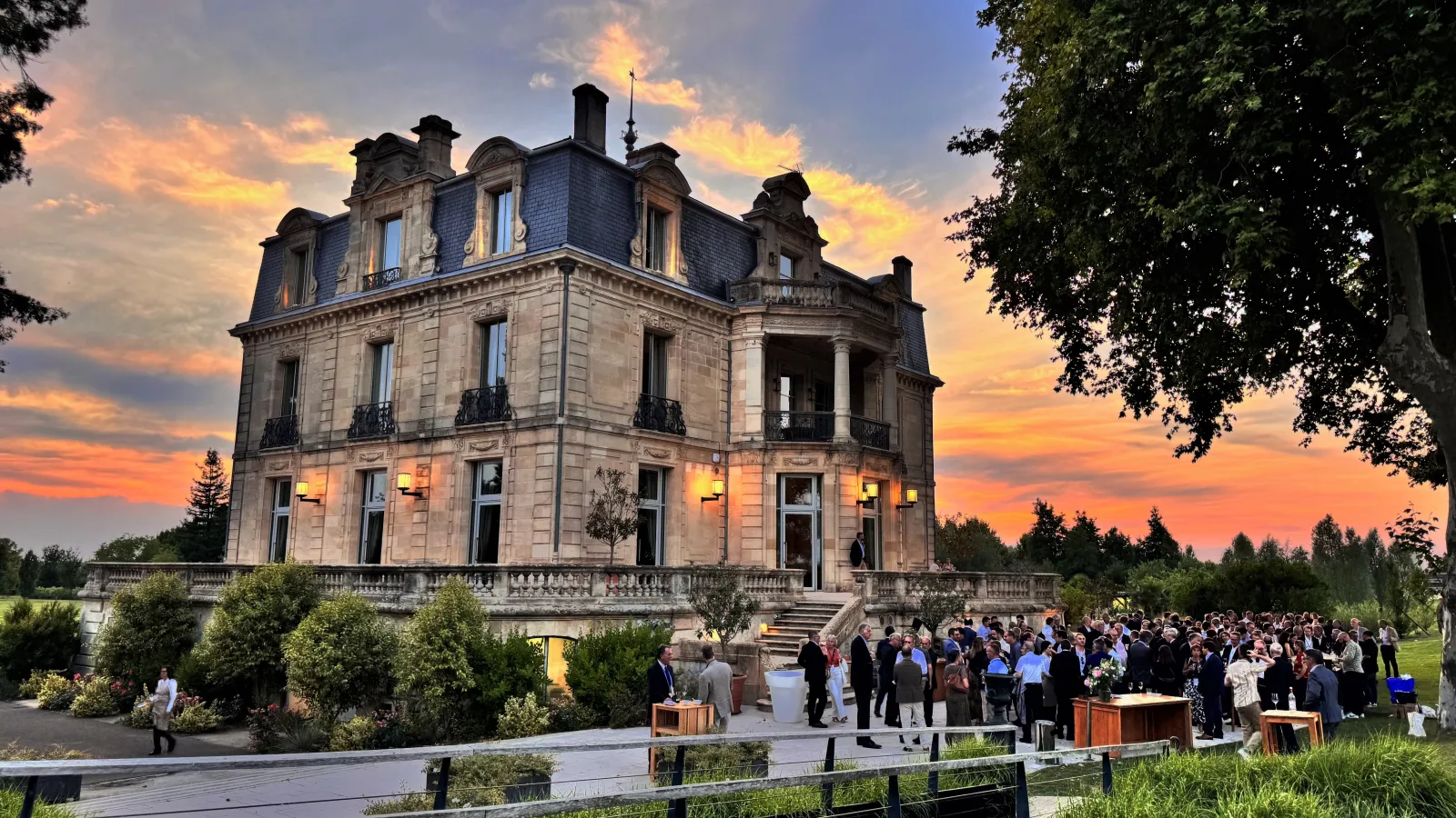 Château Grattequina in Bordeaux at sunset, a grand Second Empire mansion with mansard roof and slate turrets, surrounded by guests at an outdoor evening event, with manicured gardens and tree-lined grounds.