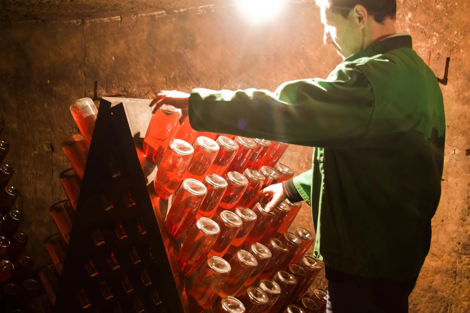 A person in a green jacket arranging red votive candles in rows inside a stone chapel or crypt, illuminated by overhead lighting.