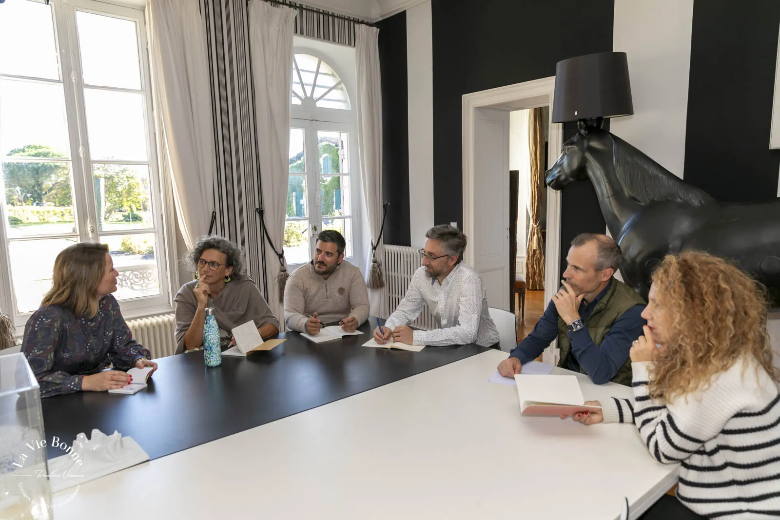 Six professionals seated around a conference table during a meeting in a modern, bright room with large windows overlooking a garden, engaged in discussion with notebooks and materials.