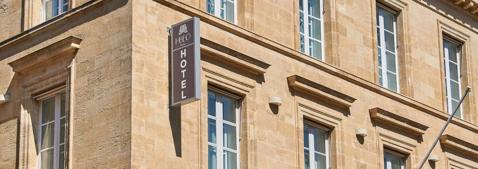 Facade of a historic stone building with a brown hotel sign reading 'HPEC HOTEL' hanging from the wall, featuring regular windows with ornate stone frames and architectural details