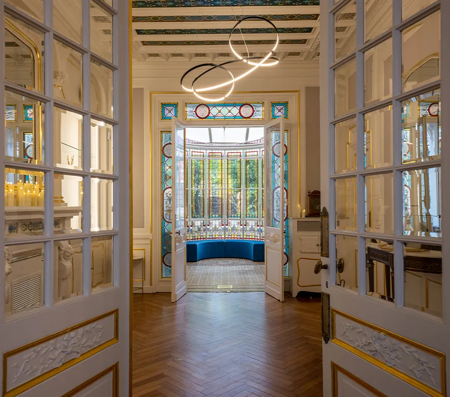 Elegant hallway with ornate double doors opening onto a luxurious room featuring a blue velvet bench, decorative stained glass windows, parquet flooring, and modern circular pendant lights, exemplifying refined Bordeaux interior design.