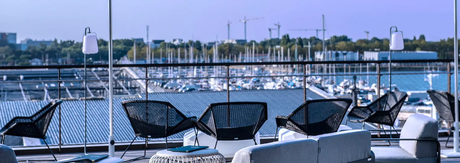 Rooftop terrace of Radisson Blu Hotel Bordeaux with black lounge chairs, white pendant lamps, and panoramic views of the Garonne River and marina with moored sailboats in the background.