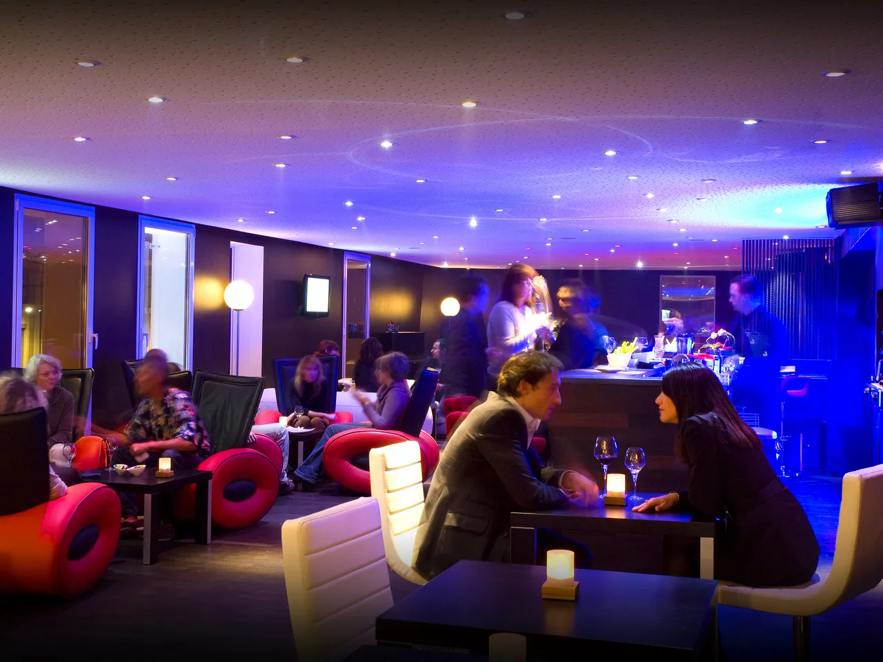 Modern lounge bar at Seeko'o Hotel Bordeaux with purple ambient lighting, red circular seating, illuminated furniture, and guests socializing at tables and the bar counter