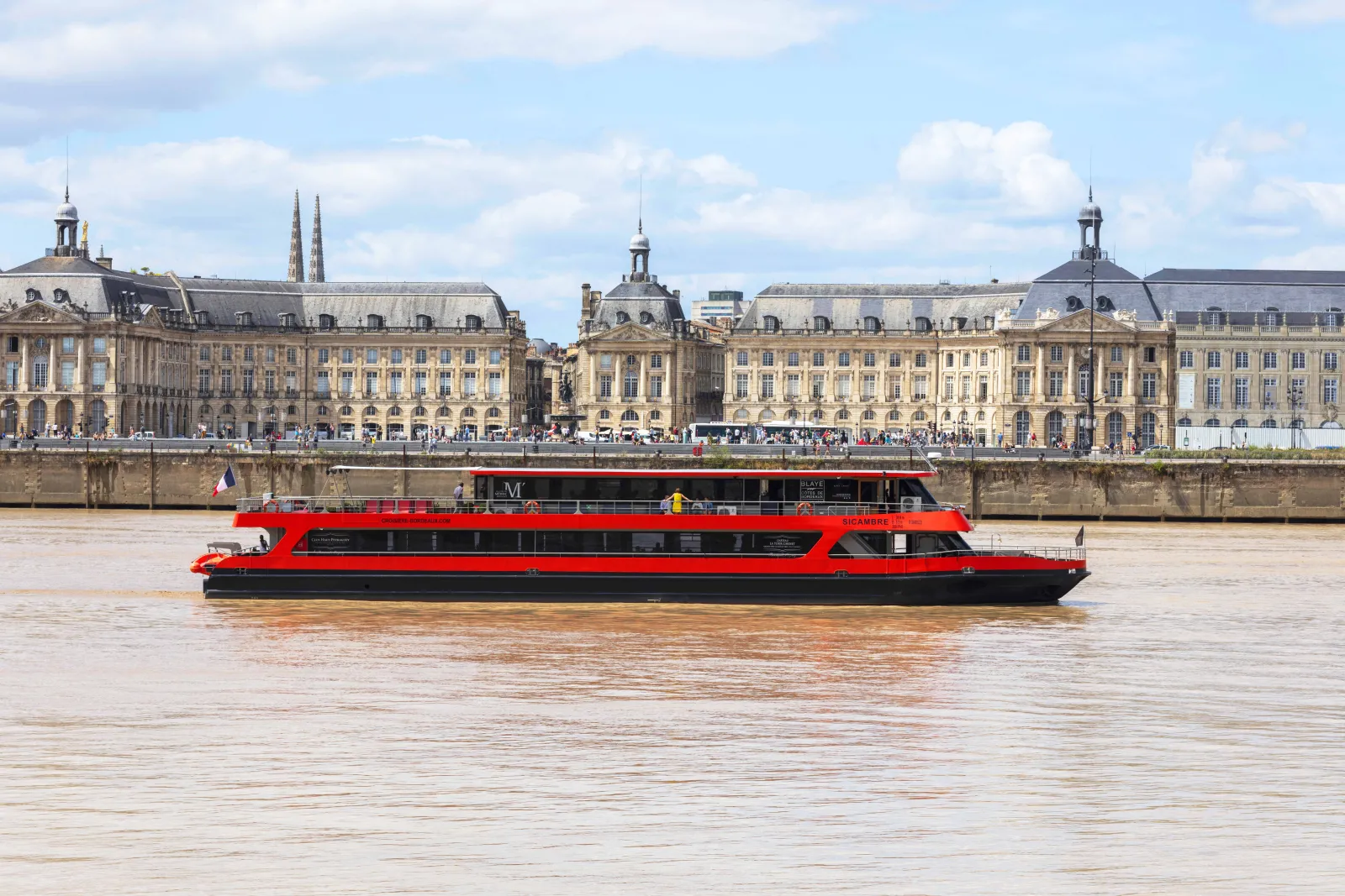 A red and black passenger riverboat cruises on the Garonne River in front of historic Belle Époque buildings in Bordeaux, France.