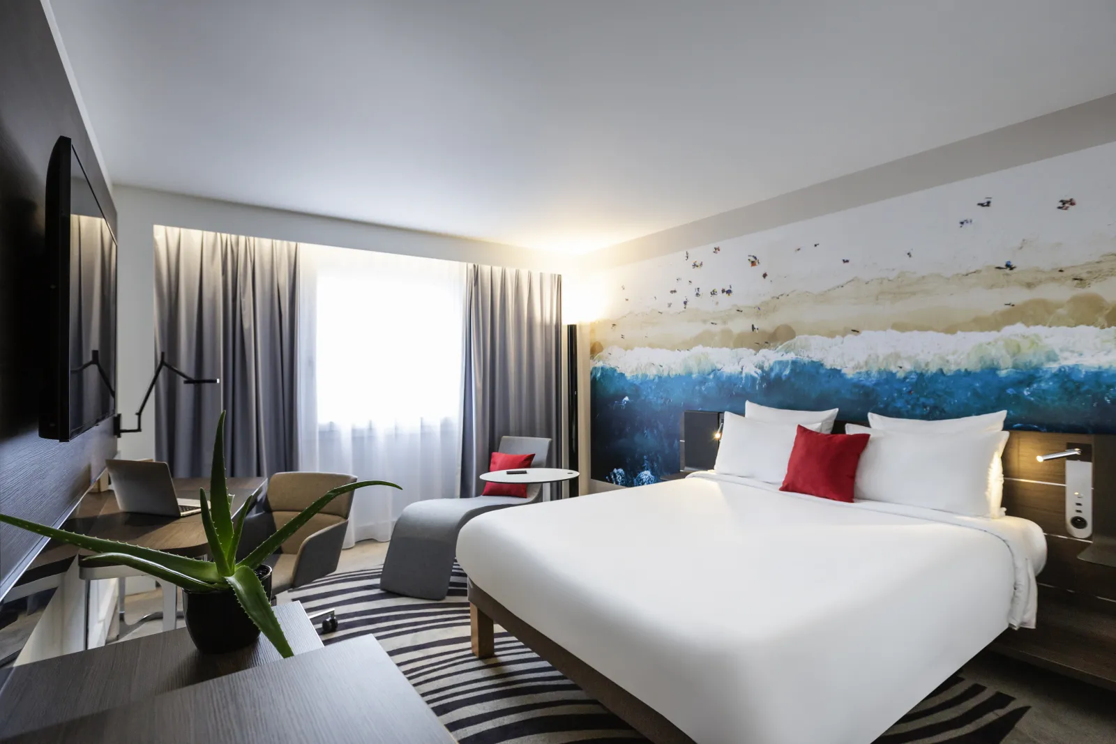 Modern hotel room at Novotel Bordeaux Centre featuring a king bed with white linens and red accent pillow, large window with curtains, wall mural of a forest landscape, desk area with laptop, flat-screen TV, and contemporary furnishings.