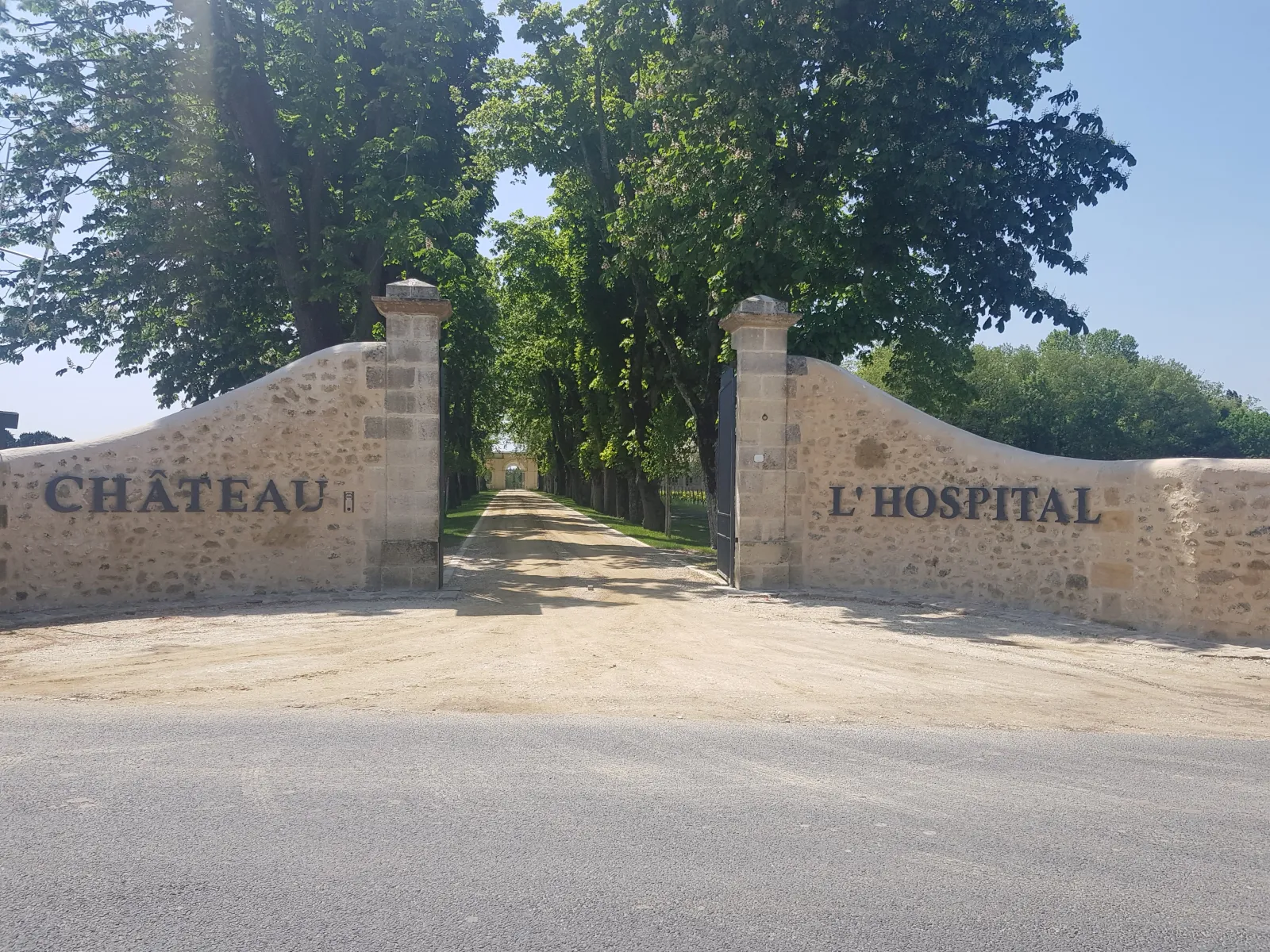 Stone gateway entrance to Château de l'Hospital in Bordeaux, with 'CHÂTEAU' and 'L'HOSPITAL' inscribed on pillars, tree-lined driveway leading to the estate
