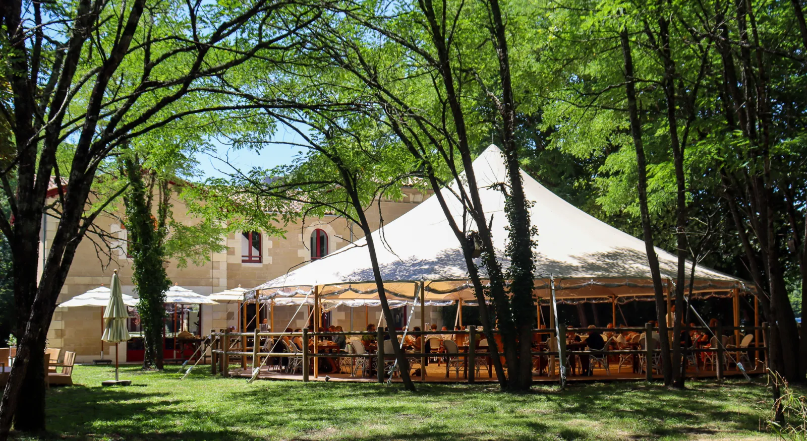 Large white tent with wooden seating area set up in a tree-lined garden next to a stone chapel building at Château Léognan in Bordeaux