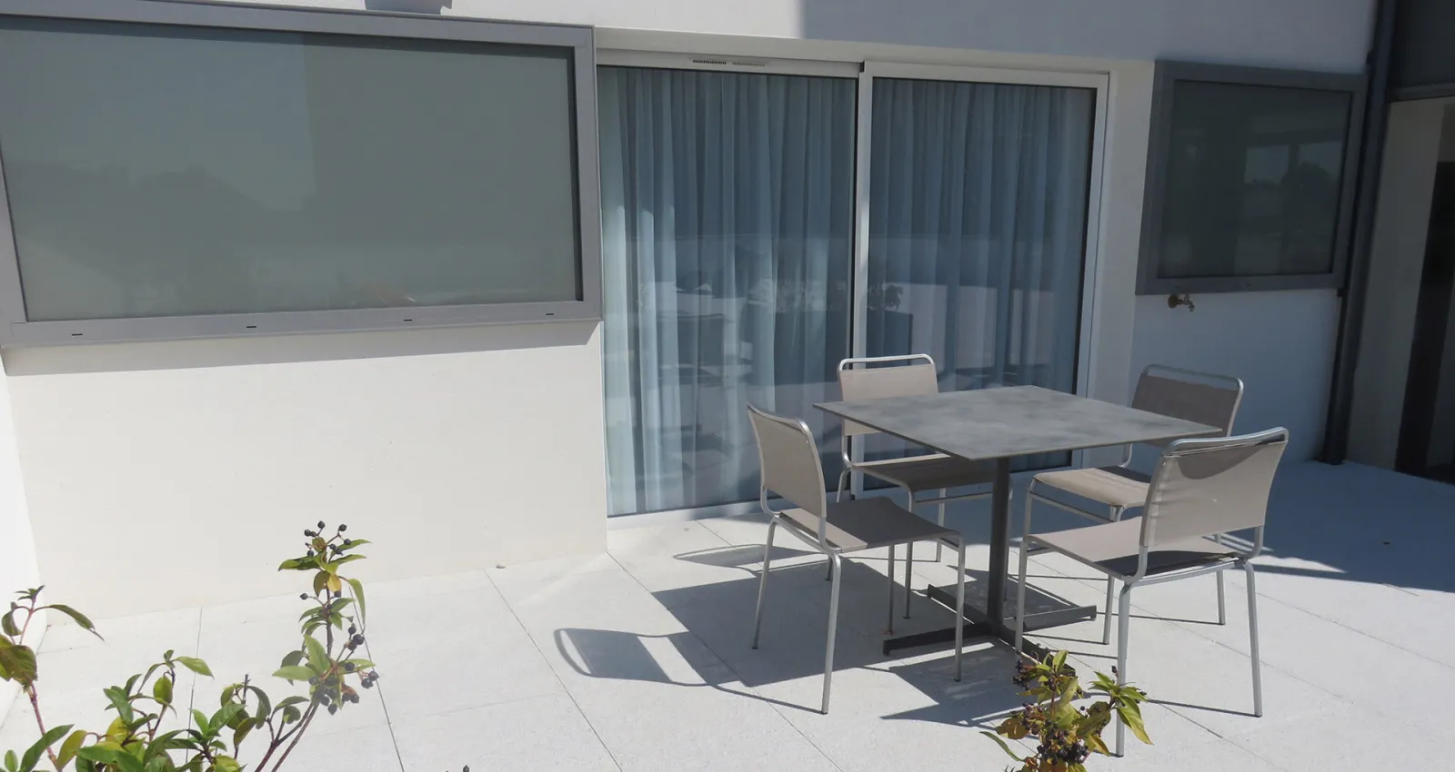 Modern terrace with dining table and chairs overlooking vertical blinds and glass doors, featuring minimalist white walls and potted plants in bright daylight.