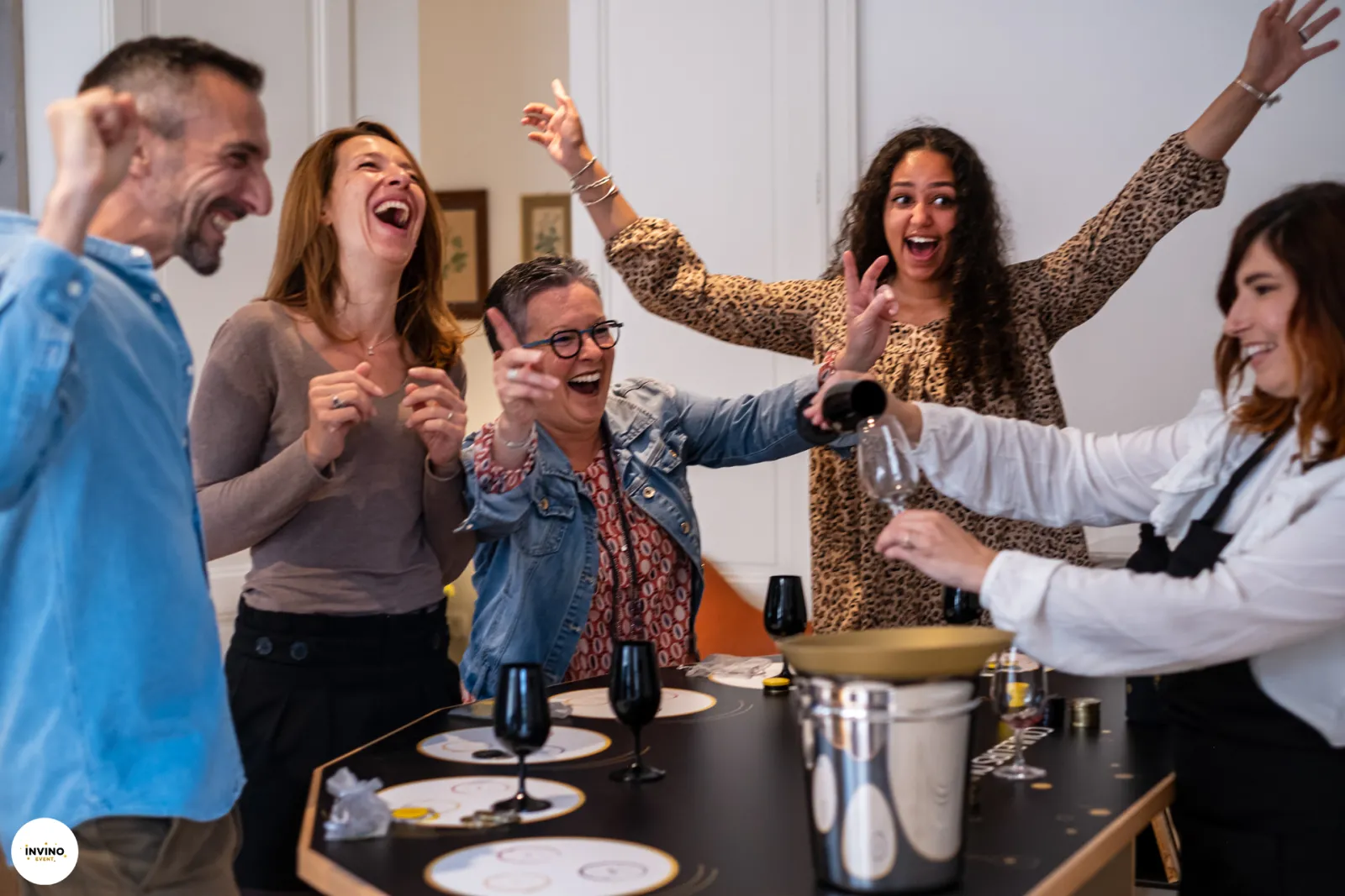 A group of five people laughing and celebrating around a dining table with wine glasses and place settings during an Invino wine event in Bordeaux.