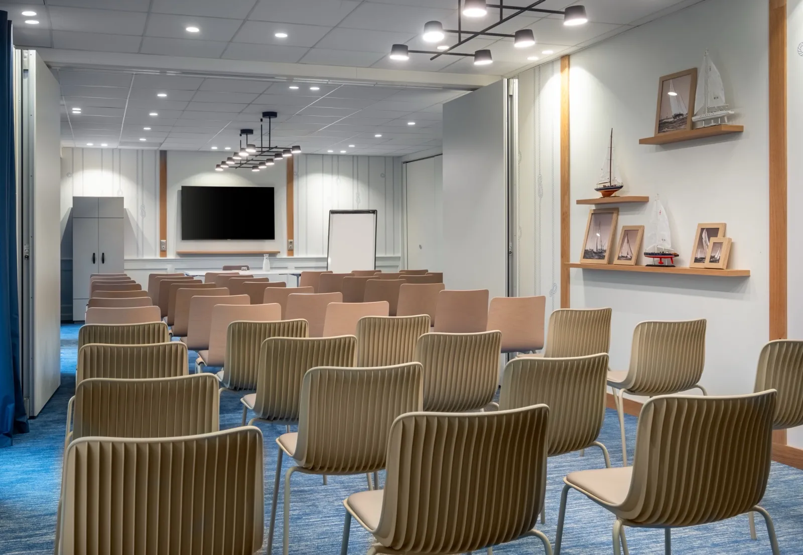 Modern conference room with rows of green striped chairs, a wall-mounted TV screen, wooden shelves with decorative sailboat models, and nautical-themed décor in a bright, contemporary space.