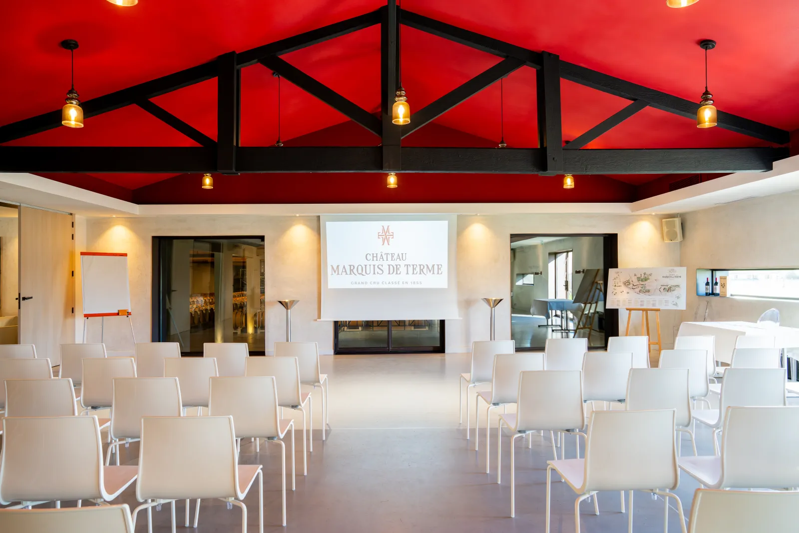 Modern conference room with red ceiling and black exposed beams at Château Marquis de Terme in Bordeaux, featuring white chairs arranged in rows facing a projection screen displaying the château's logo.