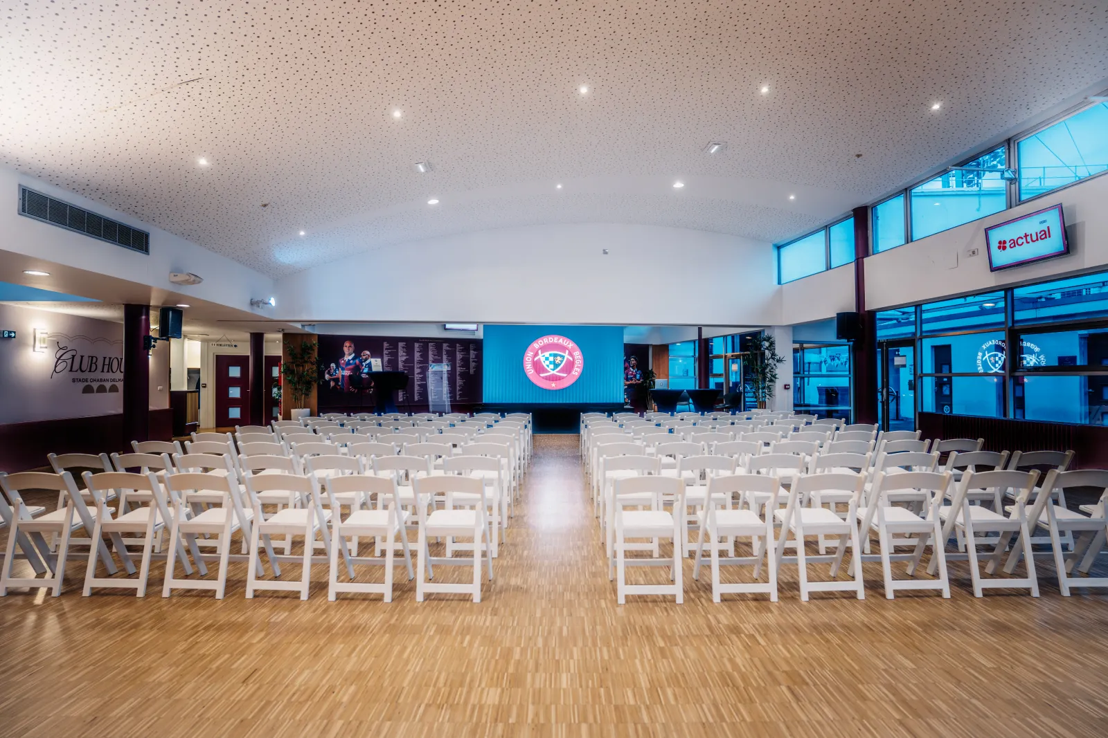 Large event space with rows of white folding chairs arranged in an auditorium setting, featuring a stage with LED screen displaying Union Bordeaux Bègles logo, modern ceiling with recessed lighting, and glass windows overlooking the city.