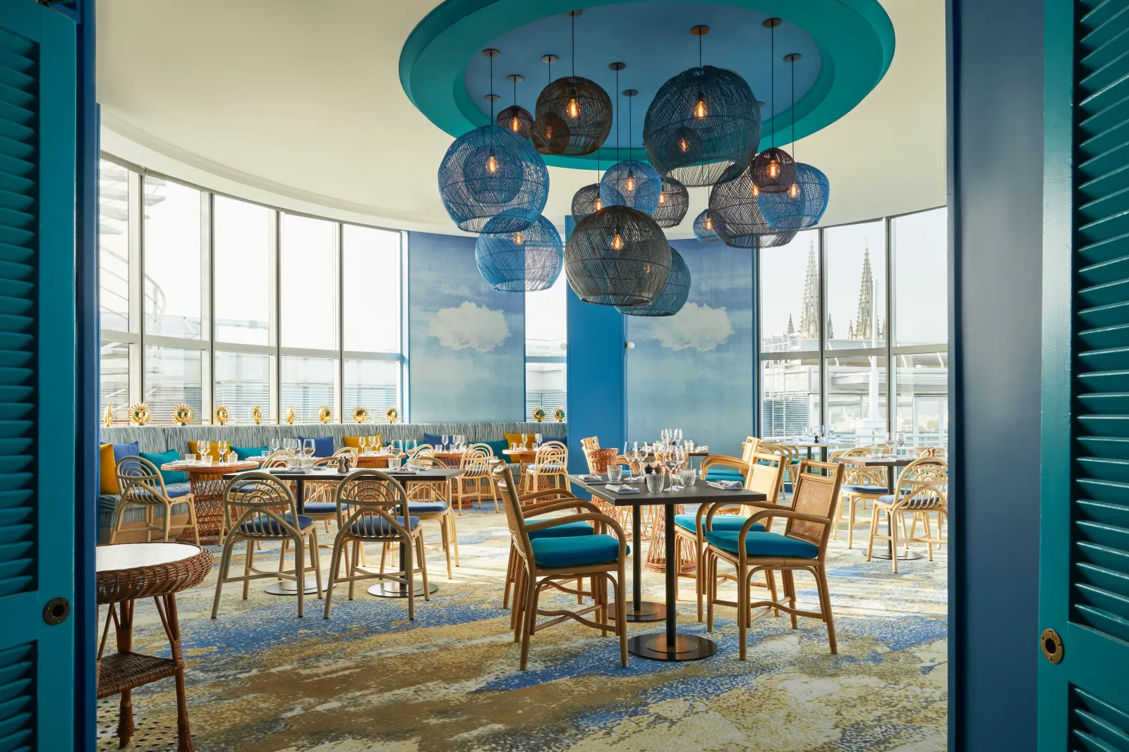 Modern restaurant interior at Indigo Hotel Bordeaux with turquoise ceiling, suspended blue and bronze mesh pendant lights, wooden chairs with blue cushions, large windows overlooking the city, and contemporary furnishings.