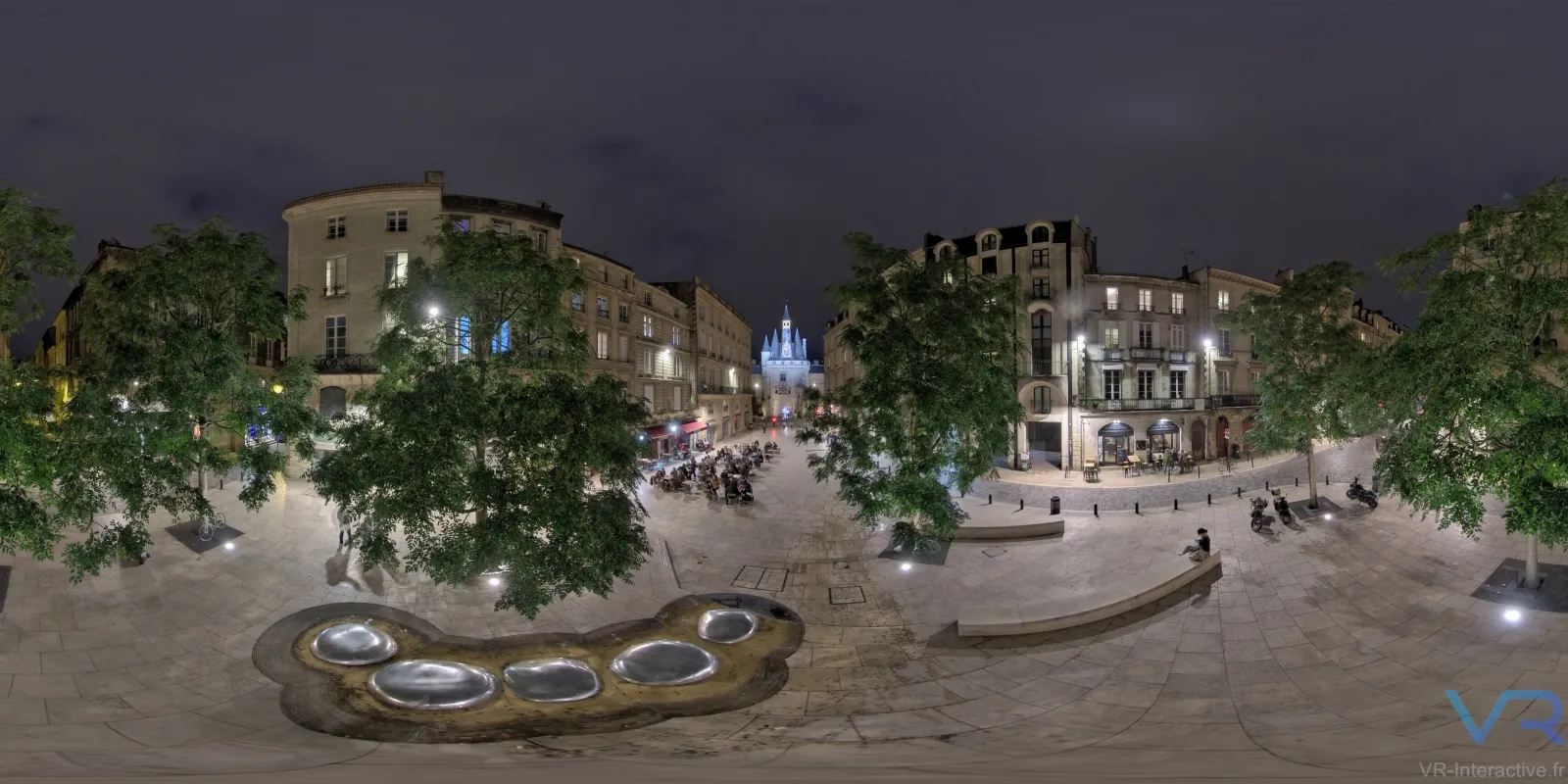 Night view of a Bordeaux city square with historic Haussmann-style buildings, illuminated trees, decorative ground sculptures, and a distant lit church spire, rendered as a 360-degree virtual reality scene.
