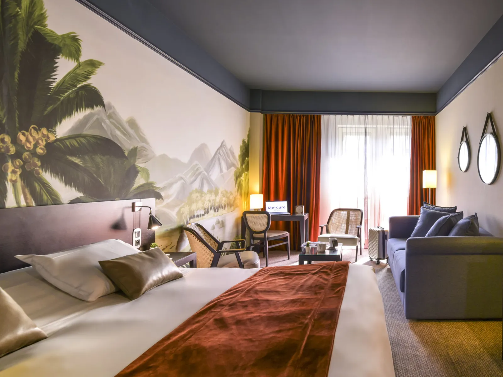 Modern hotel room at Mercure Bordeaux Château Chartrons featuring a bed with rust-colored runner, work desk, seating area, large mural of sunflowers and mountains, orange curtains, and contemporary décor