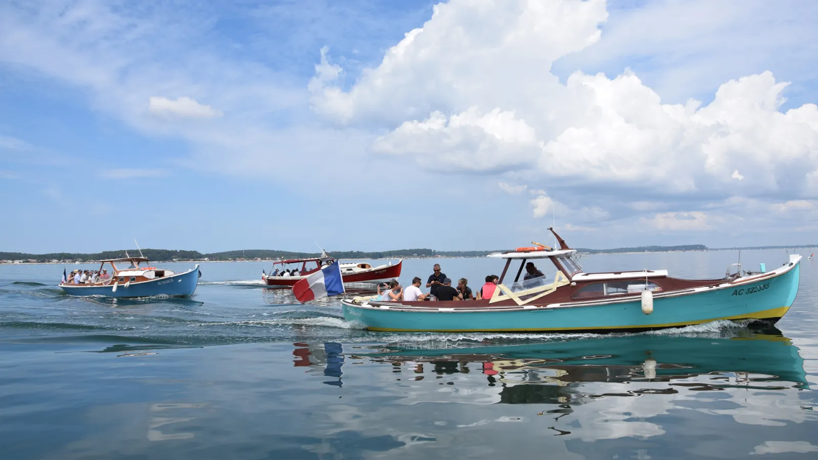 Several colorful fishing boats moored in calm blue waters near Bordeaux, with white clouds and green coastline in the background.