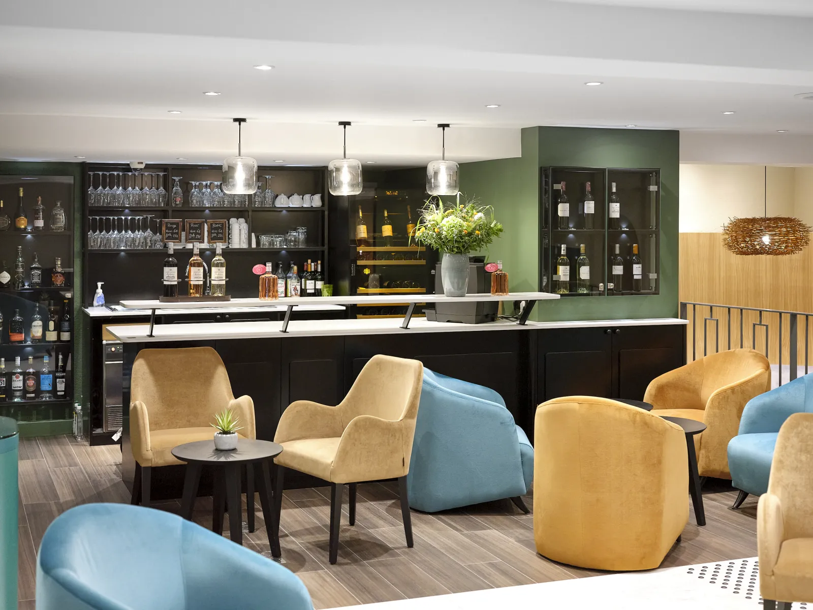Modern hotel lounge with a black bar counter, green accent wall, pendant lights, and comfortable seating featuring mustard and blue armchairs arranged around small tables. Shelving displays spirits and glasses behind the bar.