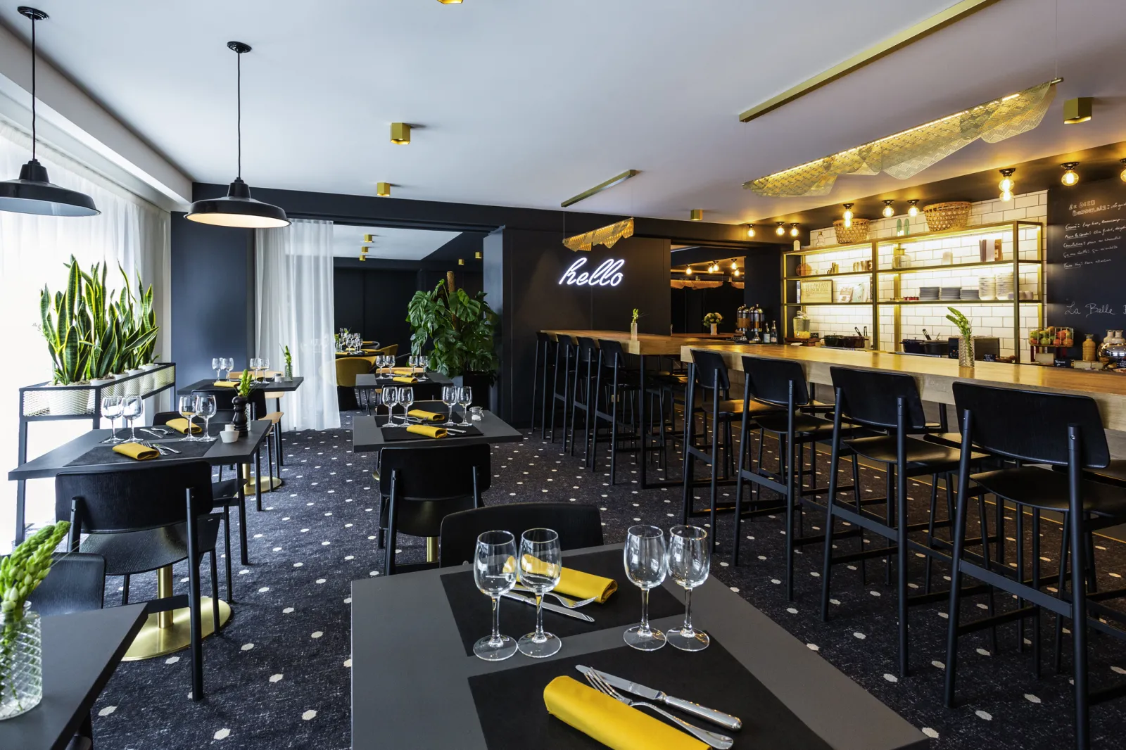 Modern restaurant and bar interior with black tables set with yellow napkins and glassware, a long wooden bar with black stools, illuminated shelving, neon 'hello' sign, pendant lighting, and decorative plants in the Mercure Bordeaux Centre hotel.
