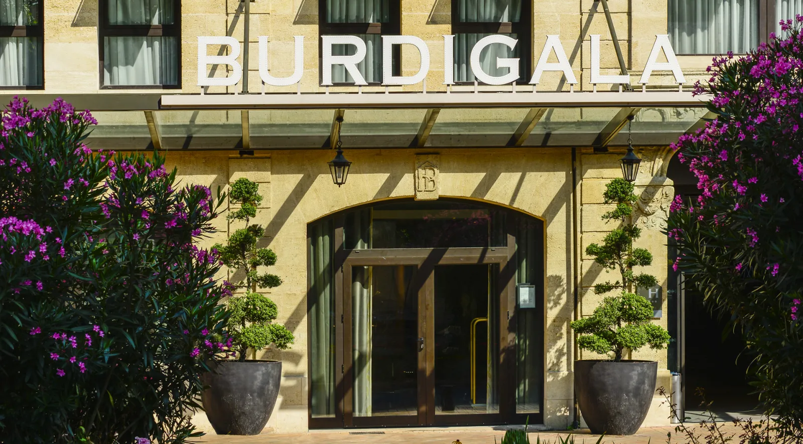 Entrance of the Burdigala Hotel in Bordeaux, featuring a stone facade with white signage, arched doorway, and vibrant purple flowers in dark planters on either side