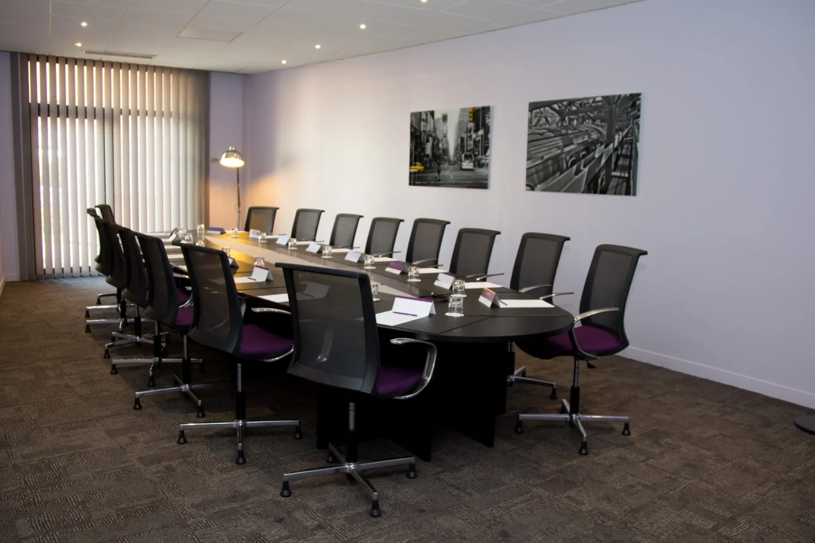 Modern conference room with a long black table, black and purple office chairs, floor-to-ceiling windows with vertical blinds, recessed ceiling lights, and black and white urban photography on white walls.