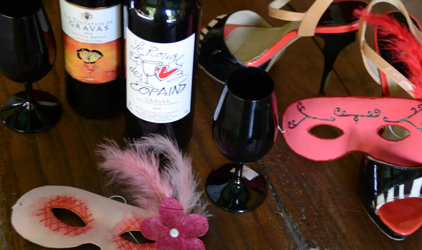 Festive party setup with Bordeaux wines, decorative masquerade masks in pink and red with feathers, ribbons, and wine glasses on a wooden table