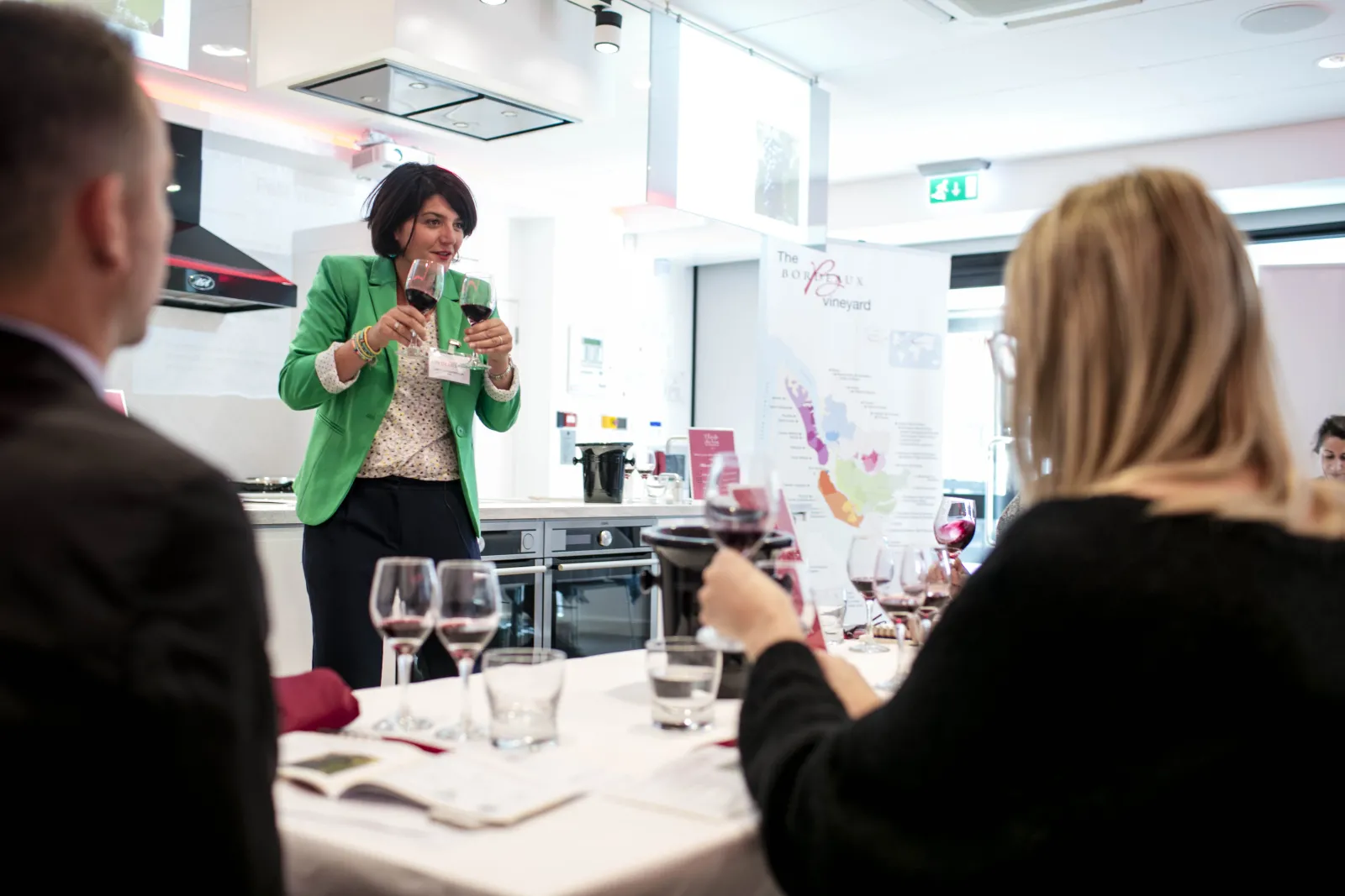 A woman in a green blazer presents at a wine tasting event in a modern kitchen, holding a microphone while speaking to seated guests with wine glasses on the table.