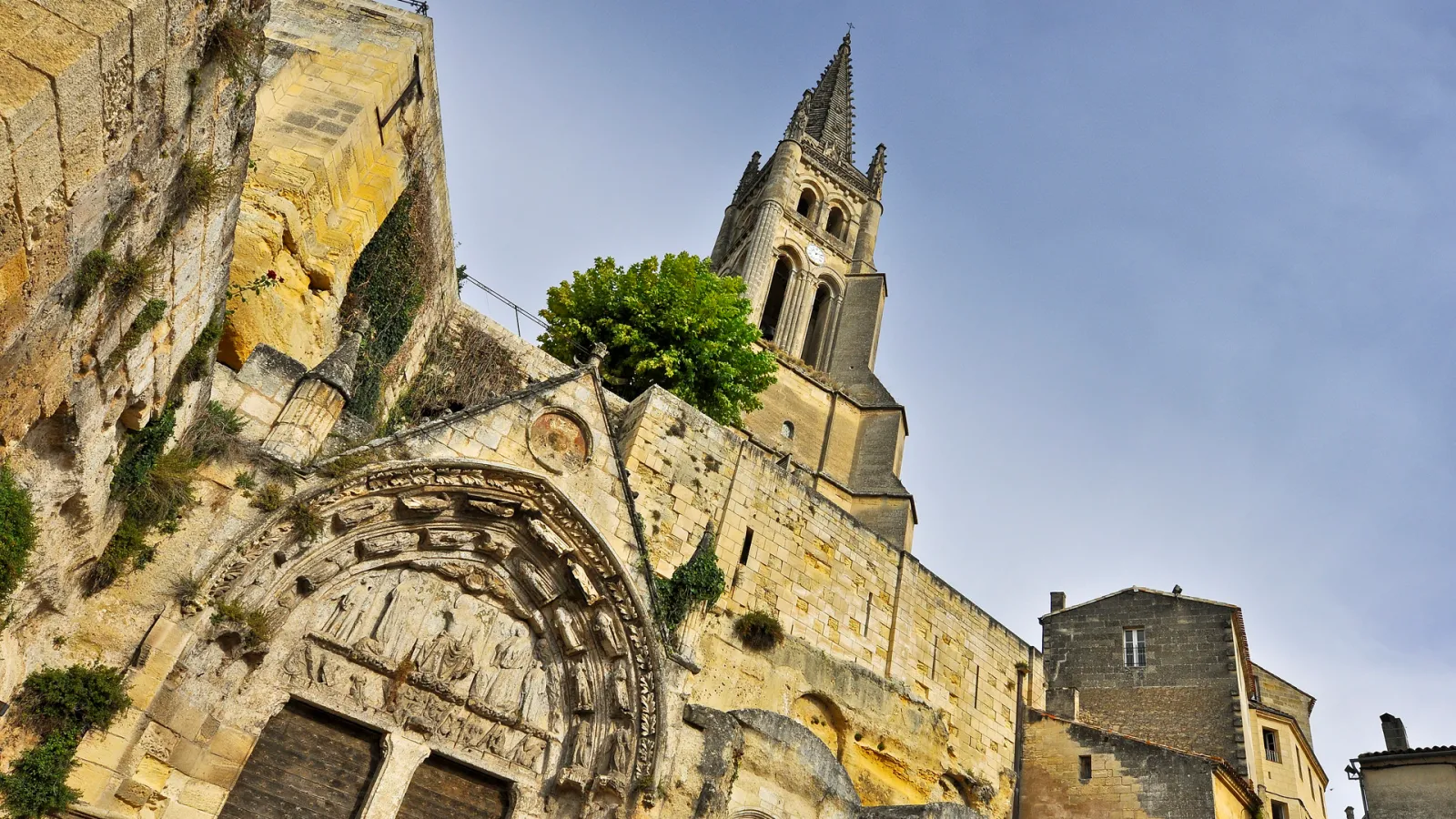 Historic stone church with ornate Romanesque portal and tall spire in Saint-Emilion, featuring weathered golden limestone facade and climbing ivy.