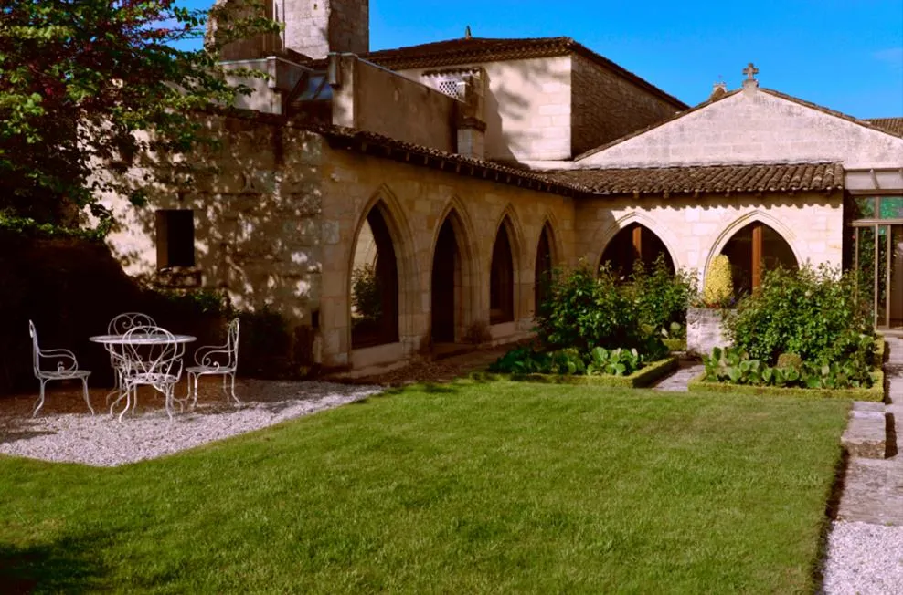 Courtyard of the Jacobins Convent in Bordeaux featuring a green lawn, gravel seating area with wrought iron furniture, Gothic arches, and ivy-covered walls under a clear blue sky.
