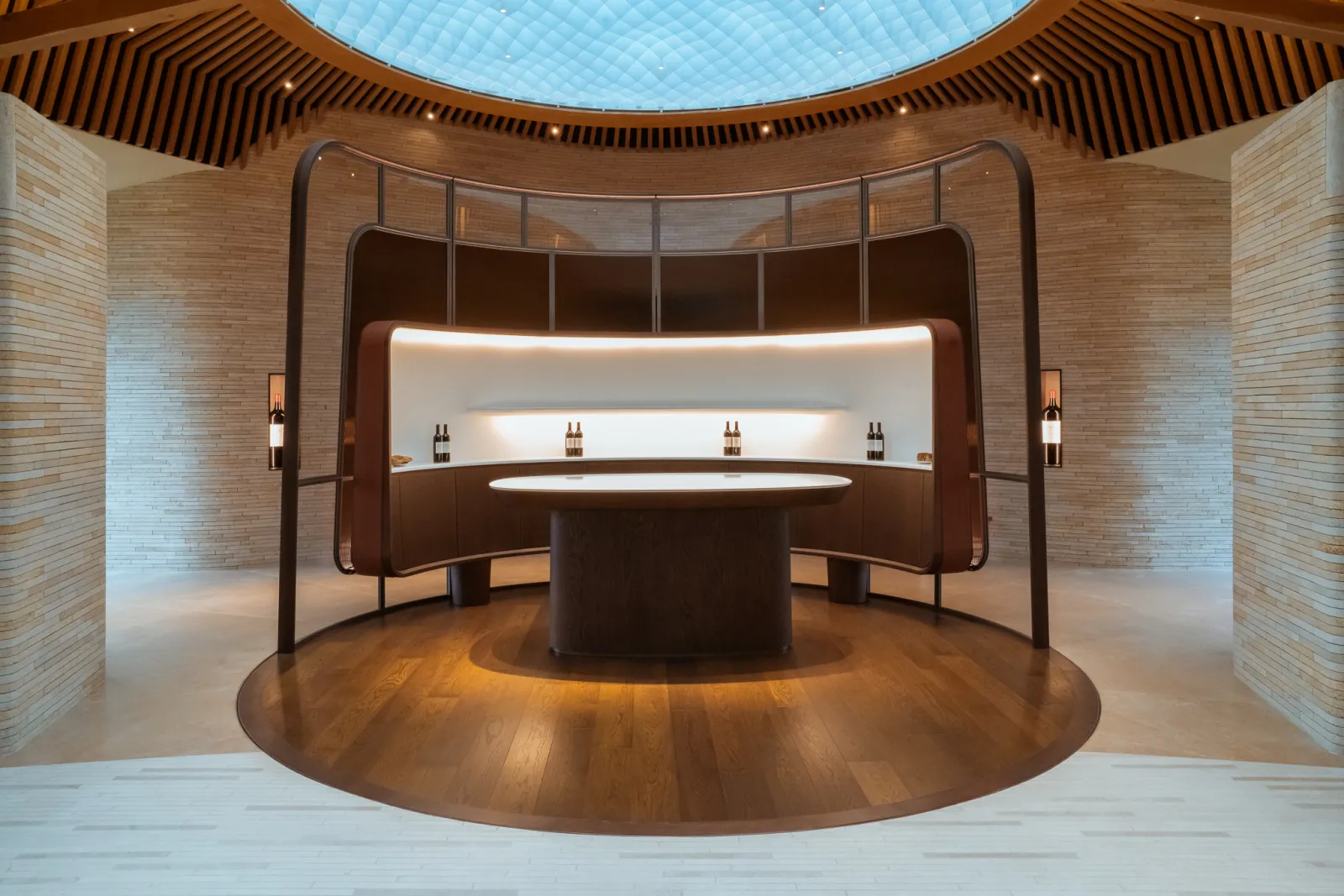 Modern circular tasting room with curved wooden counter, display shelves with wine bottles, brick walls, skylight ceiling, and wooden flooring in a contemporary architectural design