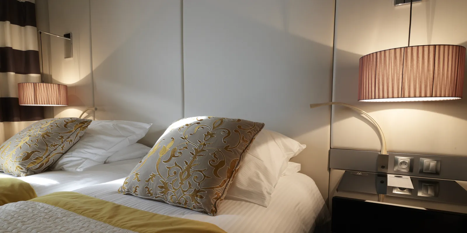 Luxury hotel bedroom at Burdigala Bordeaux featuring a queen bed with decorative gold-patterned pillows, white linens, wall-mounted pendant lights with fabric shades, and modern minimalist design with beige and gray tones.