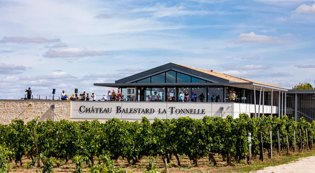 Modern winery building of Château Balestard La Tonnelle with vineyard in foreground, located in Bordeaux wine region, featuring contemporary architecture with large windows and visitors on the terrace