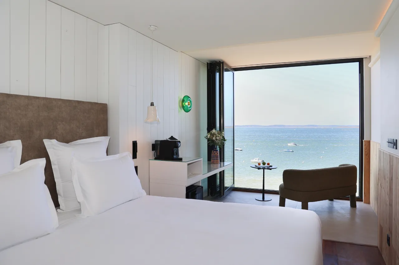 Luxury beachfront hotel bedroom with ocean view, white bed with brown headboard, modern minimalist design, floor-to-ceiling windows opening onto a Mediterranean terrace with boats visible in the water