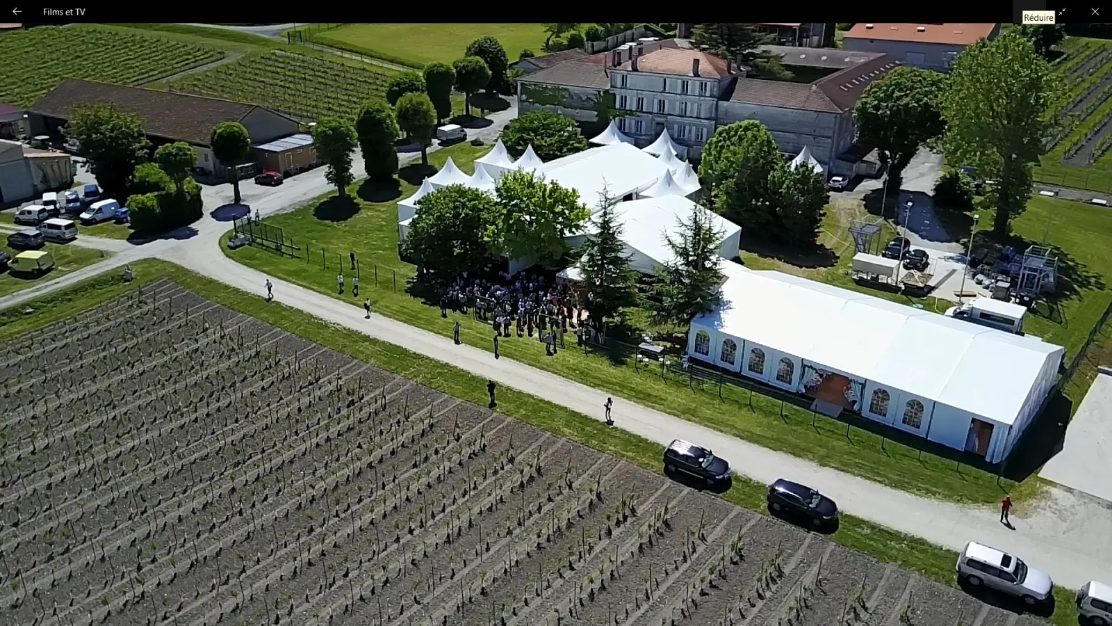 Aerial view of a wine region event setup with white marquee tents, vineyard rows, historic buildings, and parked vehicles along a pathway in a rural French landscape.