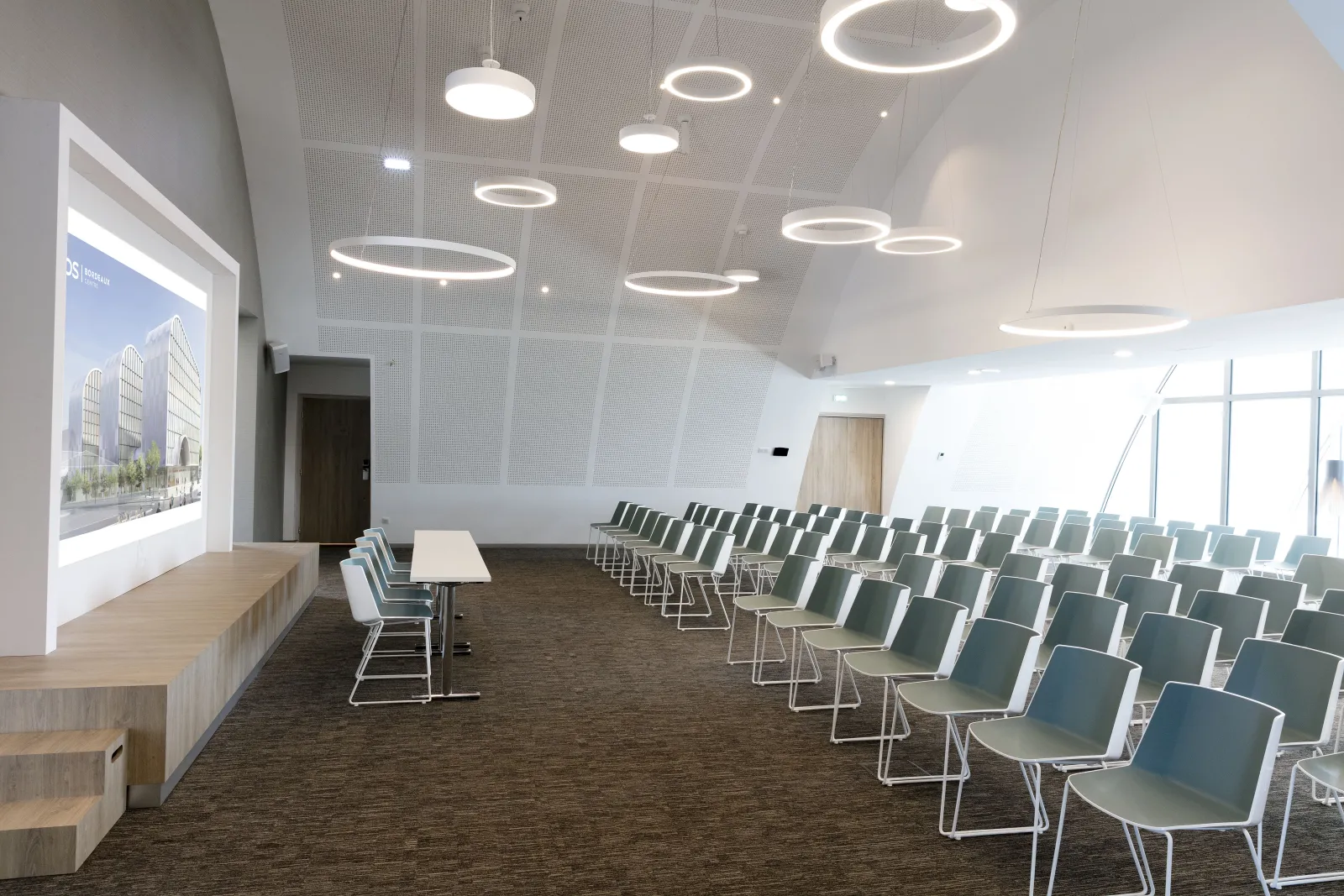 Modern conference room at Hilton Garden Inn Bordeaux Centre with rows of gray chairs, a presentation screen displaying an architectural project, wooden bench seating, and circular pendant lighting fixtures on the ceiling.