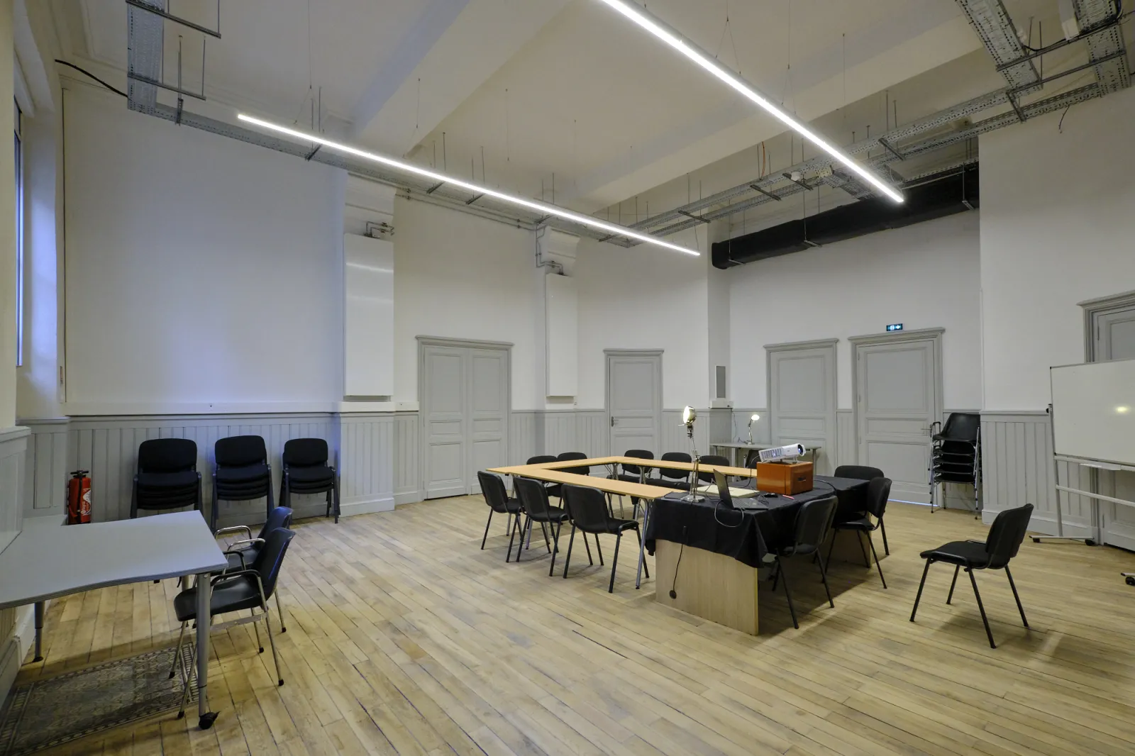 Modern event space in Bordeaux's ManuCo with wooden flooring, black chairs, wooden tables, white wainscoting, and linear ceiling lights for conferences and meetings.