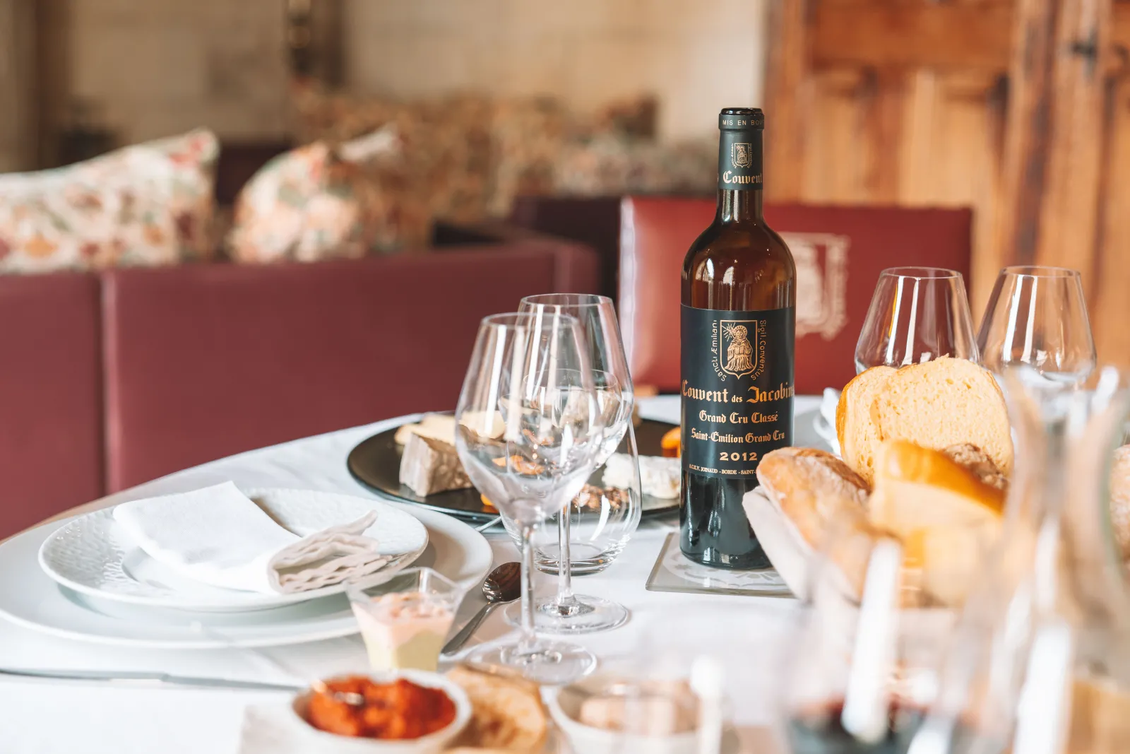 Elegant dining table setting at Couvent des Jacobins in Bordeaux, featuring fine china, wine glasses, a bottle of Bordeaux wine from 2012, fresh bread, and formal table settings in a refined interior with burgundy upholstered seating in the background.