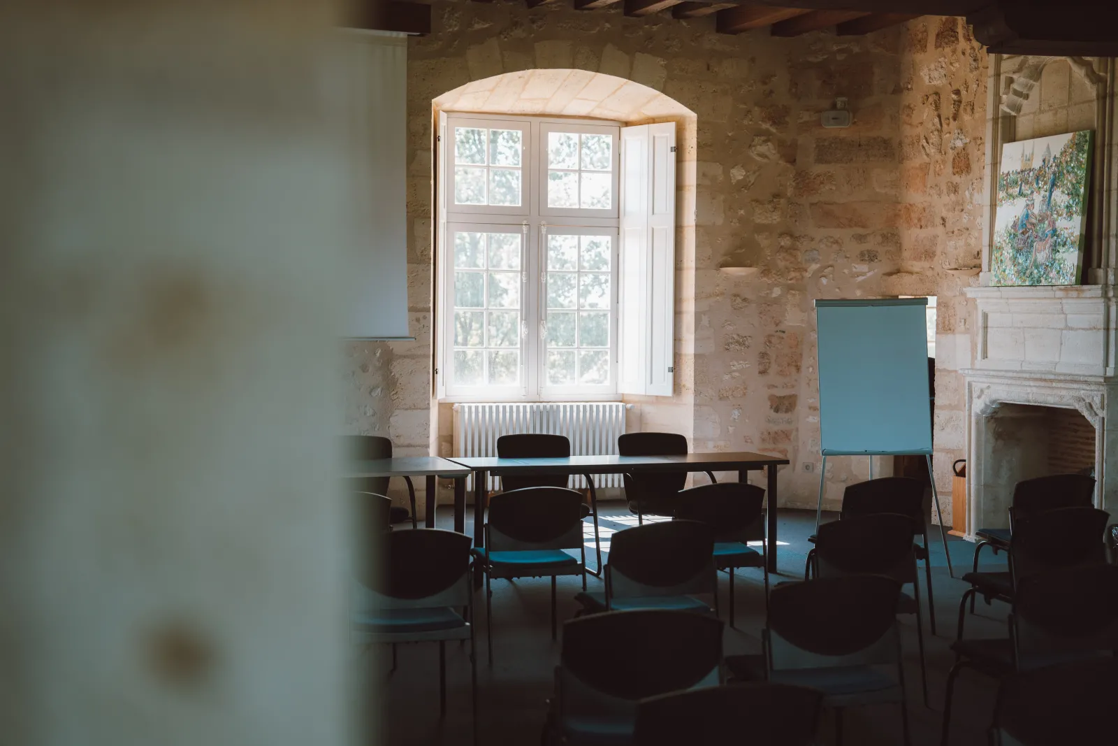 A bright meeting room with arched windows, exposed stone walls, wooden beams, black chairs arranged in a circle, and a blue flipchart, featuring natural light from tall multi-pane doors.