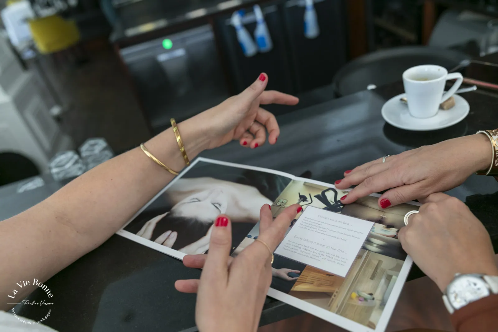 Two women with red nail polish examining photographs and documents at a table with coffee cups in Bordeaux