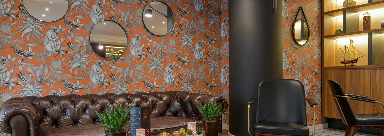 Modern hotel lounge at Mercure Bordeaux Château Chartrons featuring orange botanical wallpaper, a brown leather Chesterfield sofa, circular mirrors, black leather chairs, potted plants, and illuminated wooden shelving with decorative items.