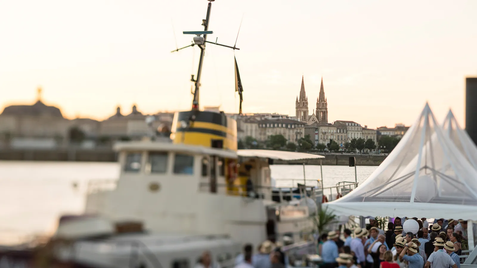 A research vessel with yellow and white hull docked at a Bordeaux waterfront event, with a white tent, crowds of visitors, and the twin spires of a cathedral visible across the river at sunset.