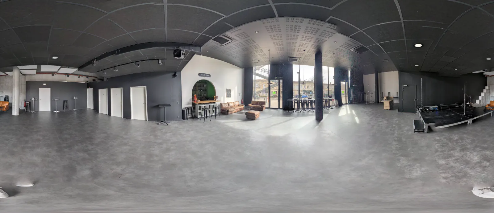 360-degree panoramic view of a large modern interior space with concrete floors, arched ceiling, support columns, and floor-to-ceiling windows showing daylight. The space includes lounge seating, a green circular architectural feature, and various zones f