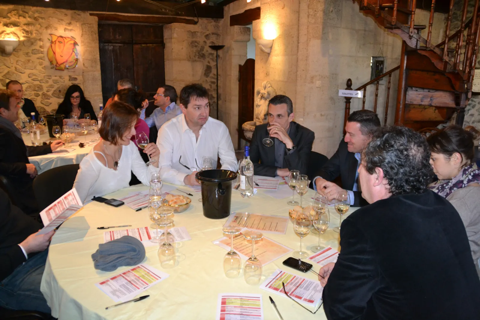 Professional business dinner in a stone-walled venue in Bordeaux, with approximately 12 people seated around a round table with white tablecloth, place settings, and colorful menu cards, discussing and networking in a formal atmosphere.