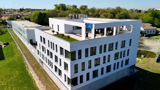 Aerial view of a modern white residential building with multiple floors and a rooftop terrace featuring blue structures, surrounded by green spaces and suburban landscape near Bordeaux.
