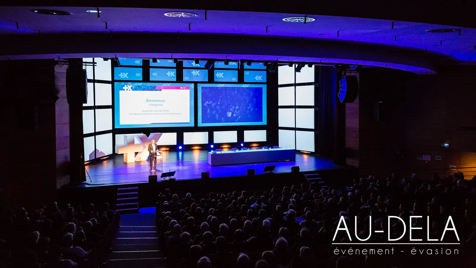 Large conference hall with a speaker on stage under blue lighting, addressing a full audience. Multiple screens display welcome message in French. Au-Delà event agency branding visible in corner.