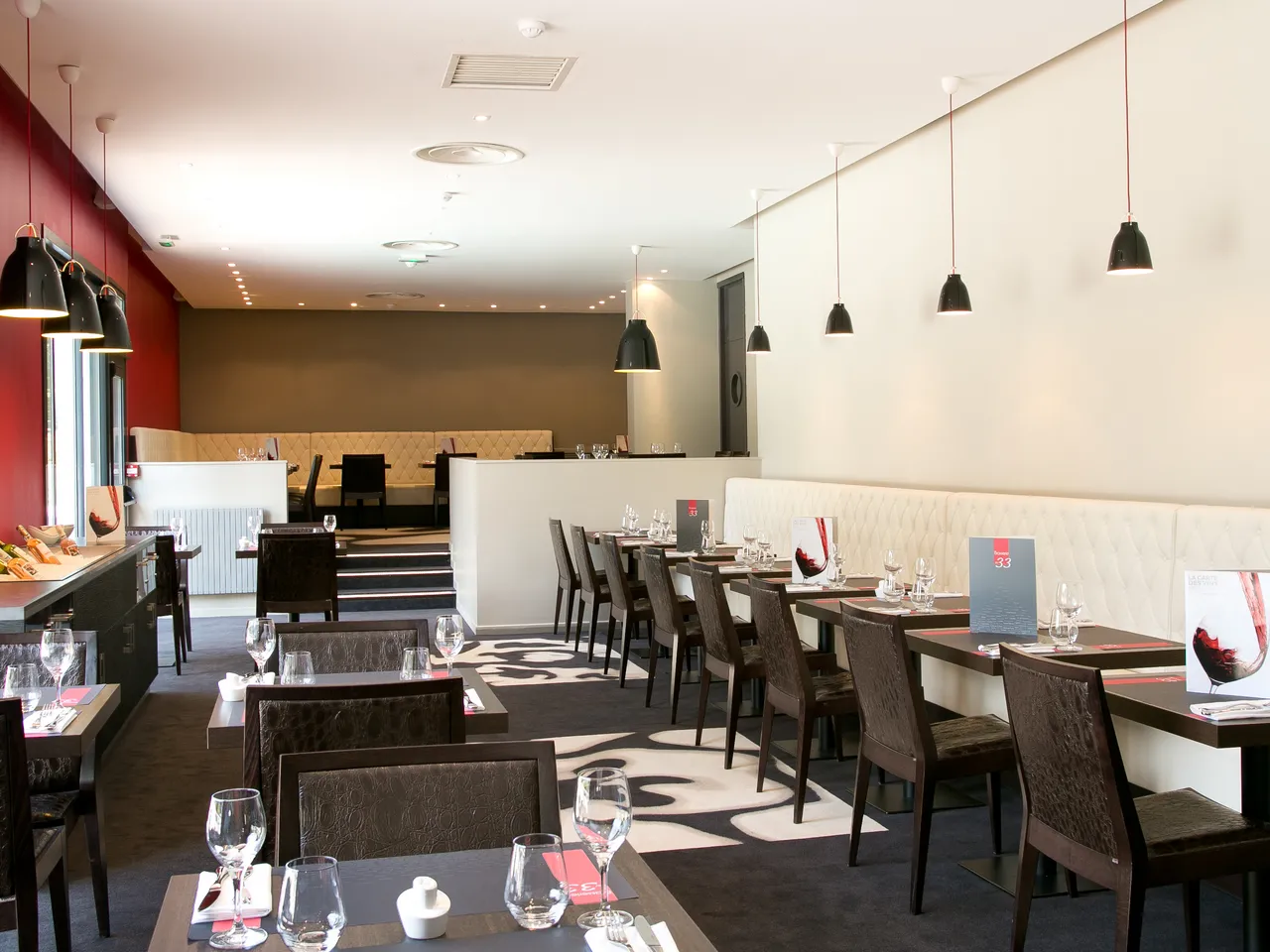 Modern restaurant dining room at Mercure Bordeaux-Lac hotel with dark tables and chairs, cream-colored banquette seating, pendant lighting, open kitchen area, and contemporary décor in red, brown, and cream tones.