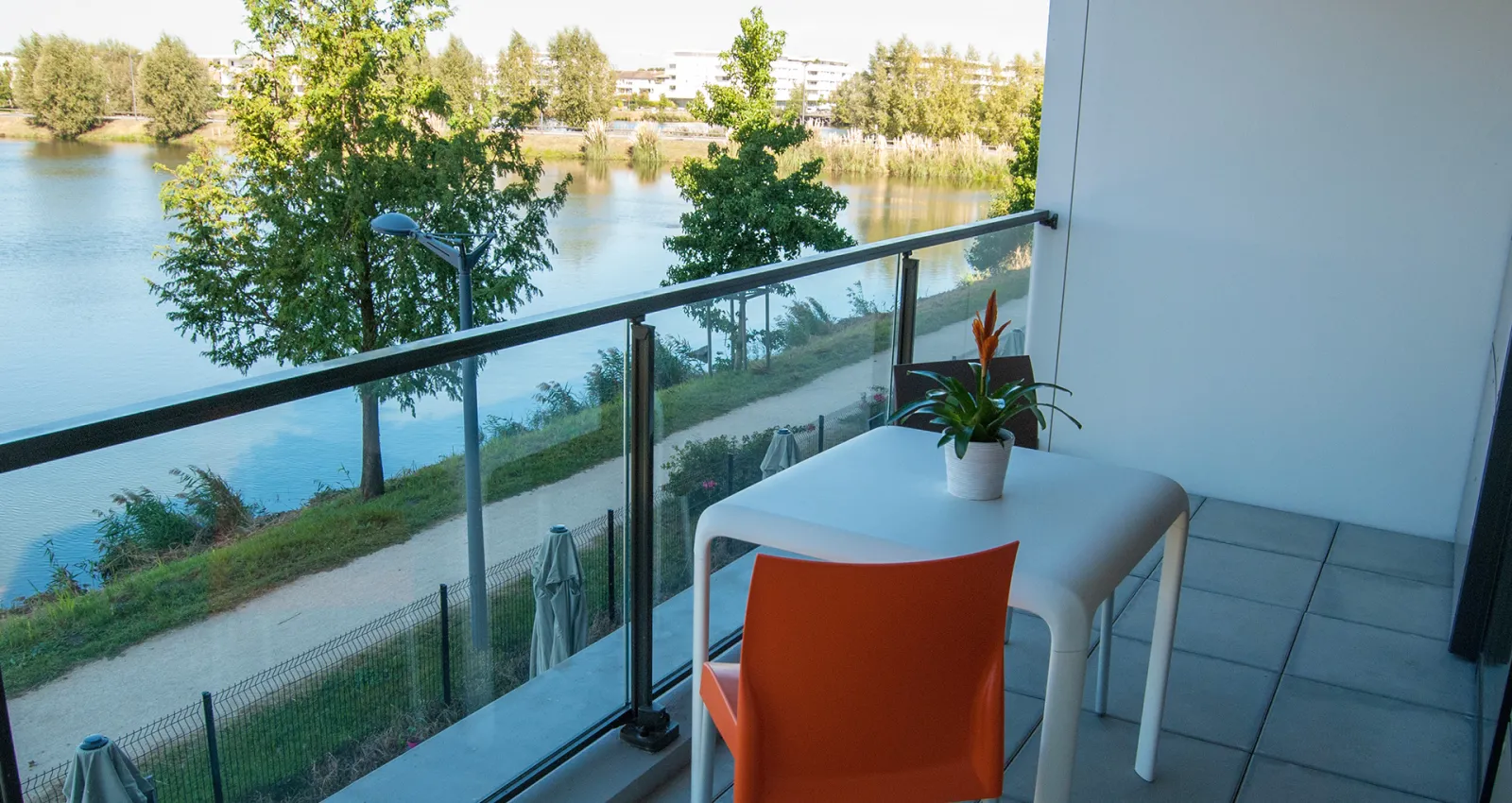 Modern balcony overlooking a calm river with green banks, trees, and residential buildings in the background. A white table with an orange chair and potted plant sit on the terrace with glass railing.