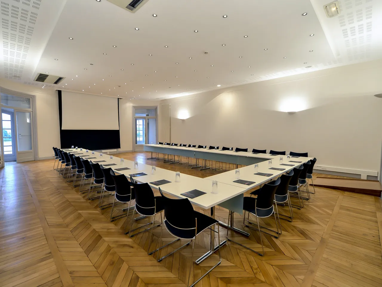 Modern conference room with U-shaped table setup, black chairs, projection screen, wooden herringbone floor, and recessed ceiling lights at Bordeaux Palais de la Bourse