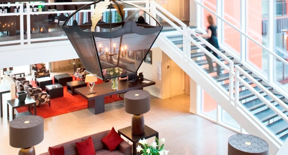 Modern luxury hotel atrium with double-height space, featuring a large sculptural pendant light, red area rug, contemporary seating areas with burgundy accents, cylindrical side tables, floor-to-ceiling windows overlooking the city, and an upper mezzanine